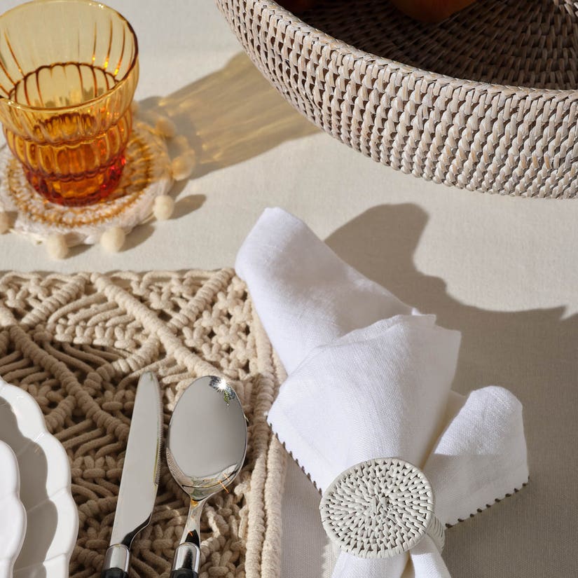 A styled tabletop setting with a white folded cloth napkin tied by a round woven napkin ring, silver cutlery on a beige crochet placemat, a translucent amber glass on a matching coaster, and a woven basket in the background.