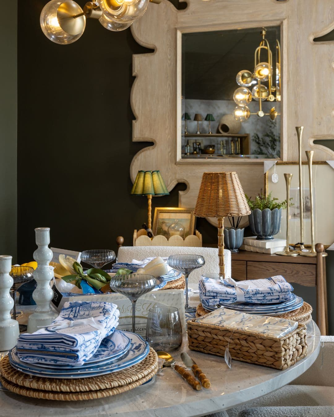 A styled dining table in a richly decorated room, featuring blue and white patterned plates, matching folded napkins, woven placemats, and a wicker basket. The background includes a dark wall, a large decorative mirror, and warm lighting from chandeliers and table lamps.