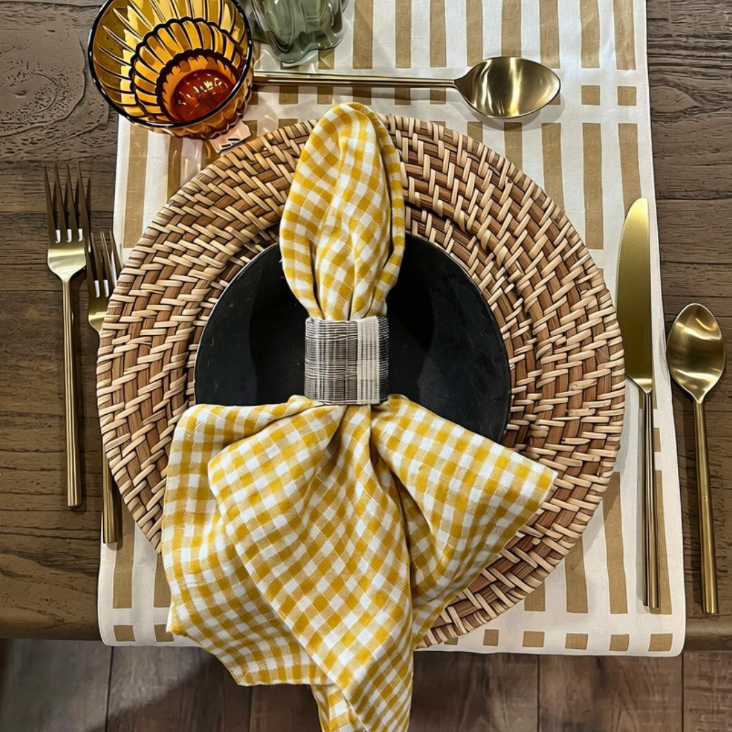 A styled tabletop place setting with a yellow and white gingham napkin tied with a plaid band, layered over a black plate and a round woven charger. A striped runner, gold-toned cutlery, a patterned glass, and a small amber drink are arranged on a wooden table.