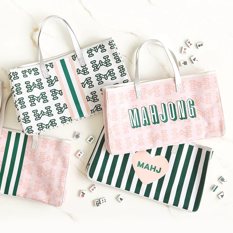 Three personalized mahjong-themed tote bags arranged on a light marble surface with scattered mahjong tiles. The bags feature green and white stripes, pink and white patterns, and the word 'MAHJONG' or a heart with 'MAHJ' printed on them.