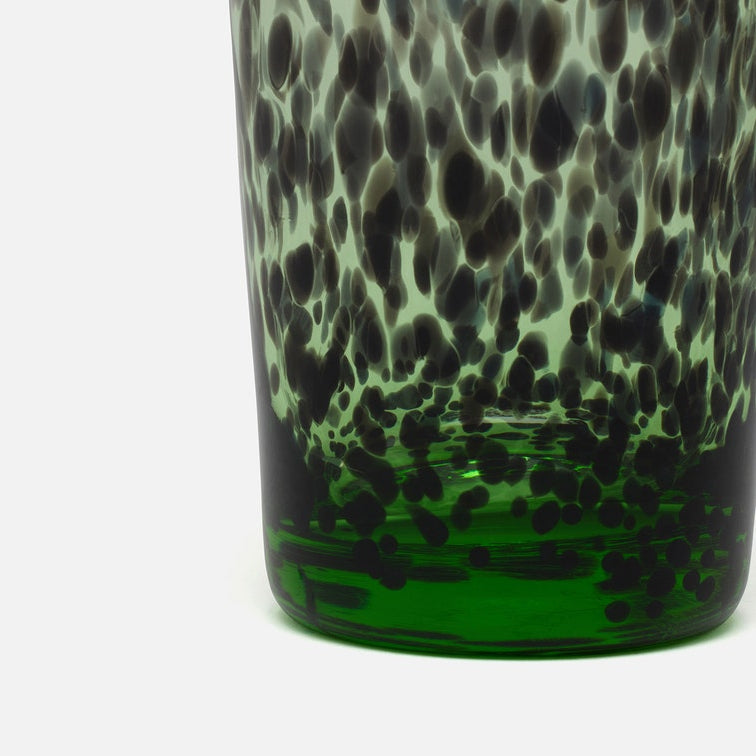 Close-up of a hand-blown glass vessel, likely a vase or candle holder, featuring a dark green base with a black and gray spotted pattern on a white background.