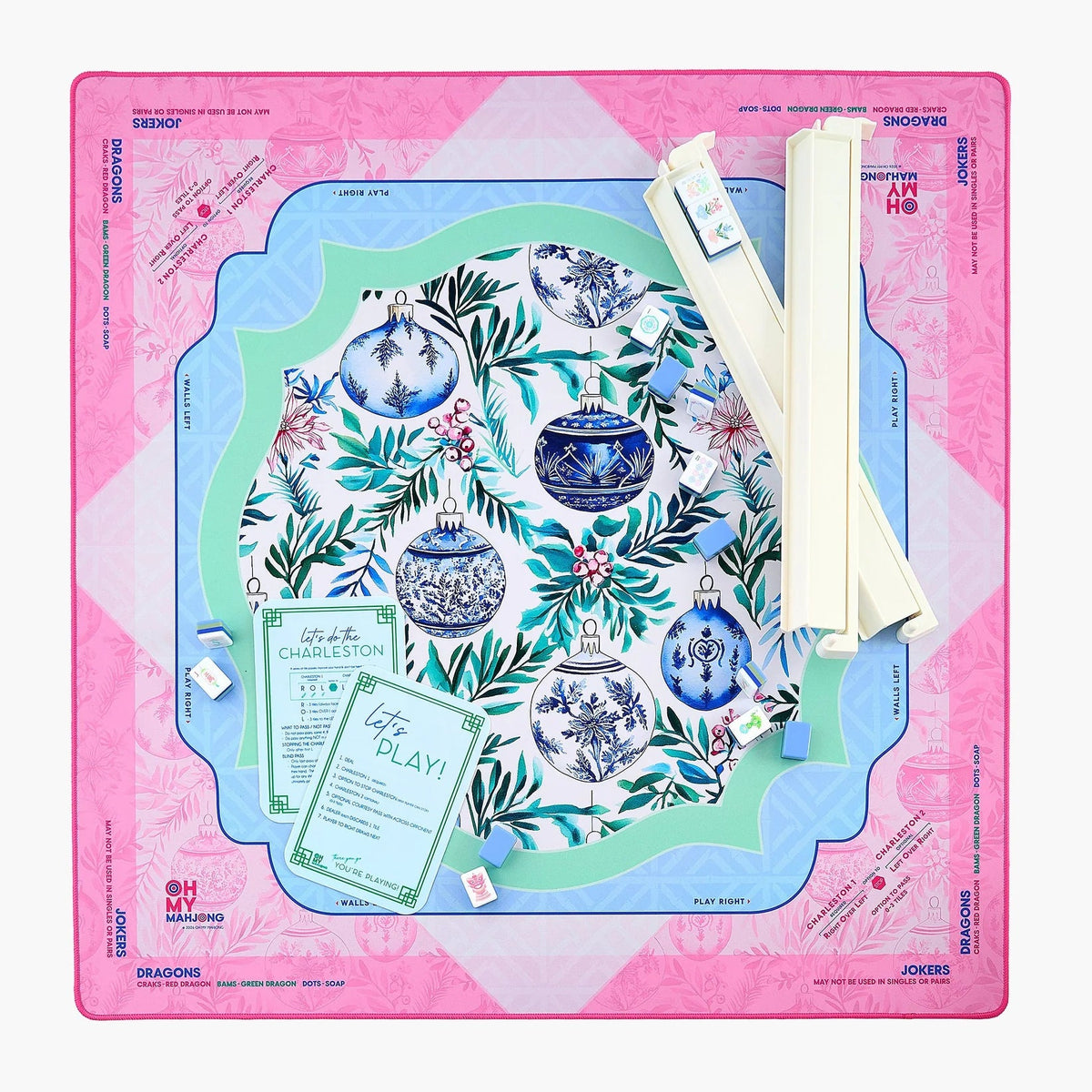 A flat-lay view of a Mahjong game set on a light gray background. The set includes a pink outer play mat with blue decorative borders, a central oval board with a blue and white floral holiday pattern, two rolled cream-colored dice, and two instruction cards.