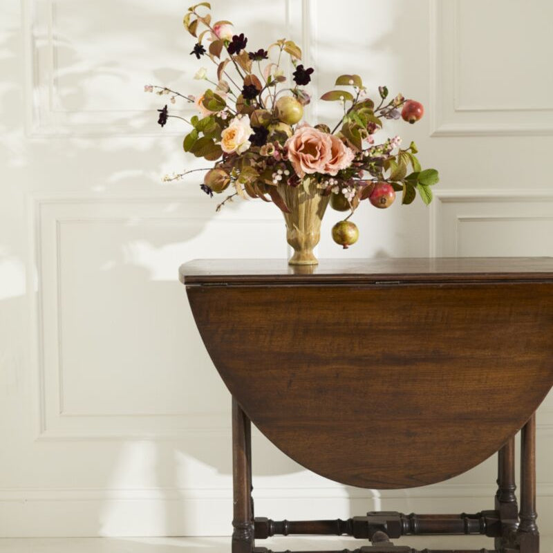 A dark brown wooden drop-leaf table with a vintage floral arrangement in a gold vase, set against a white paneled wall with soft shadows.